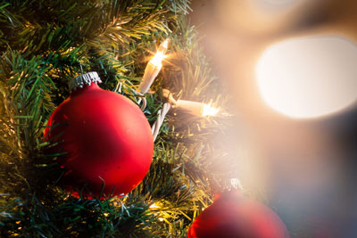 Close up of red ornament on a holiday tree. String lights are on the tree behind the ornament. There is a blurred bokeh background behind the tree.