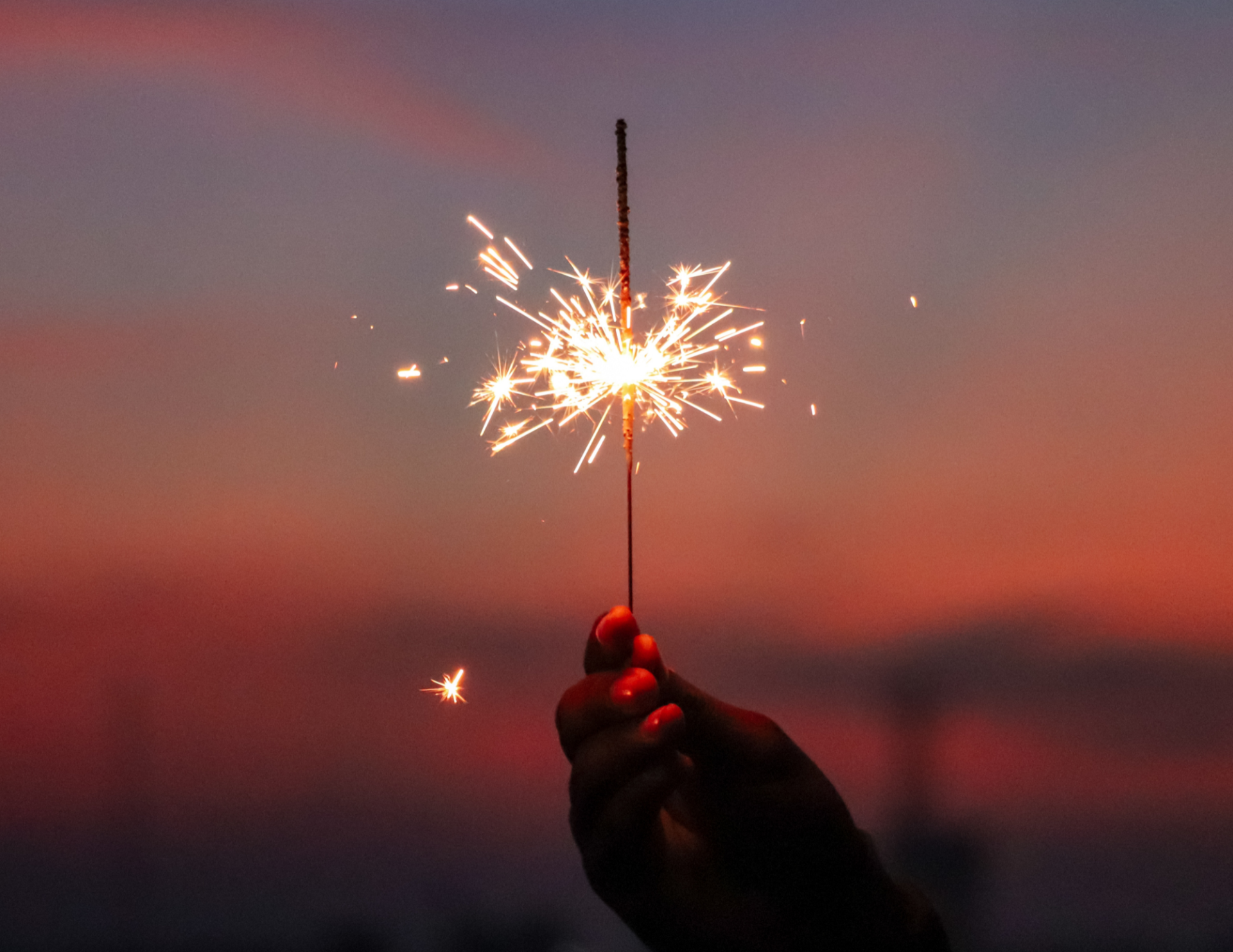 Silhouette of a hand holding a lit sparkler. The sky is at dusk in the background.