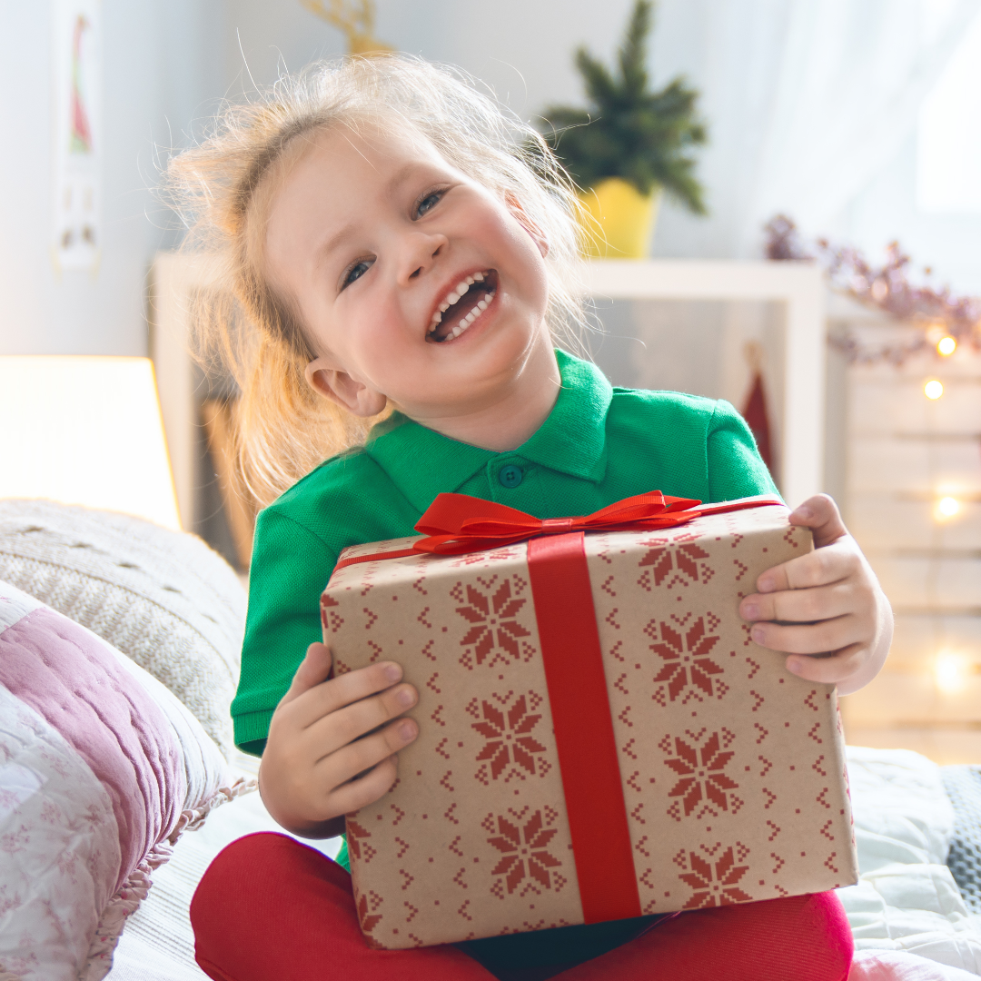 Smiling child in red and green clothing holding a present wrapped in a red bow. Child is in their room sitting on the bed. A small holiday tree is in the background. 