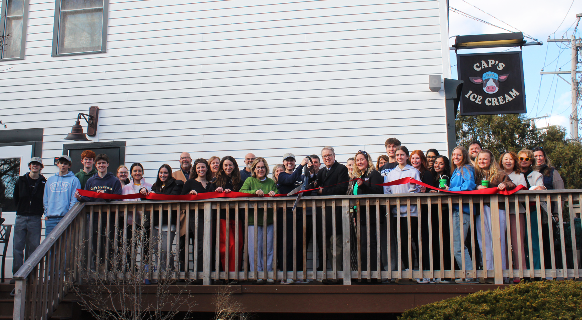Mayor Paul Hoefert, staff from Caps Ice Cream, and community members lined up in front of the Caps Ice Cream building. A red ribbon spans the line of people. The Mayor and Caps owner hold a large pair of scissors with intent to cut the ribbon.