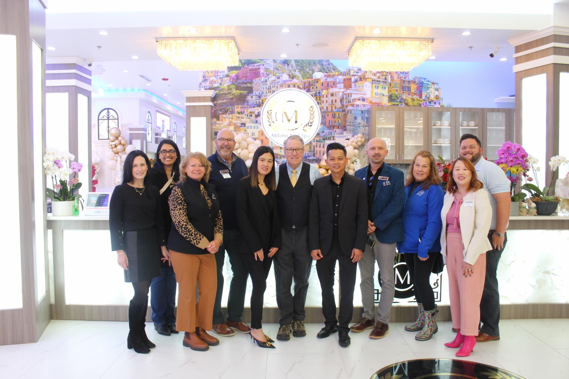 Mayor Paul Hoefert, owners of Milano Nail Spa, and community members posed together smiling. The group is in the front lobby of the business. A logo sign, lights, and elegant decor are in the background.