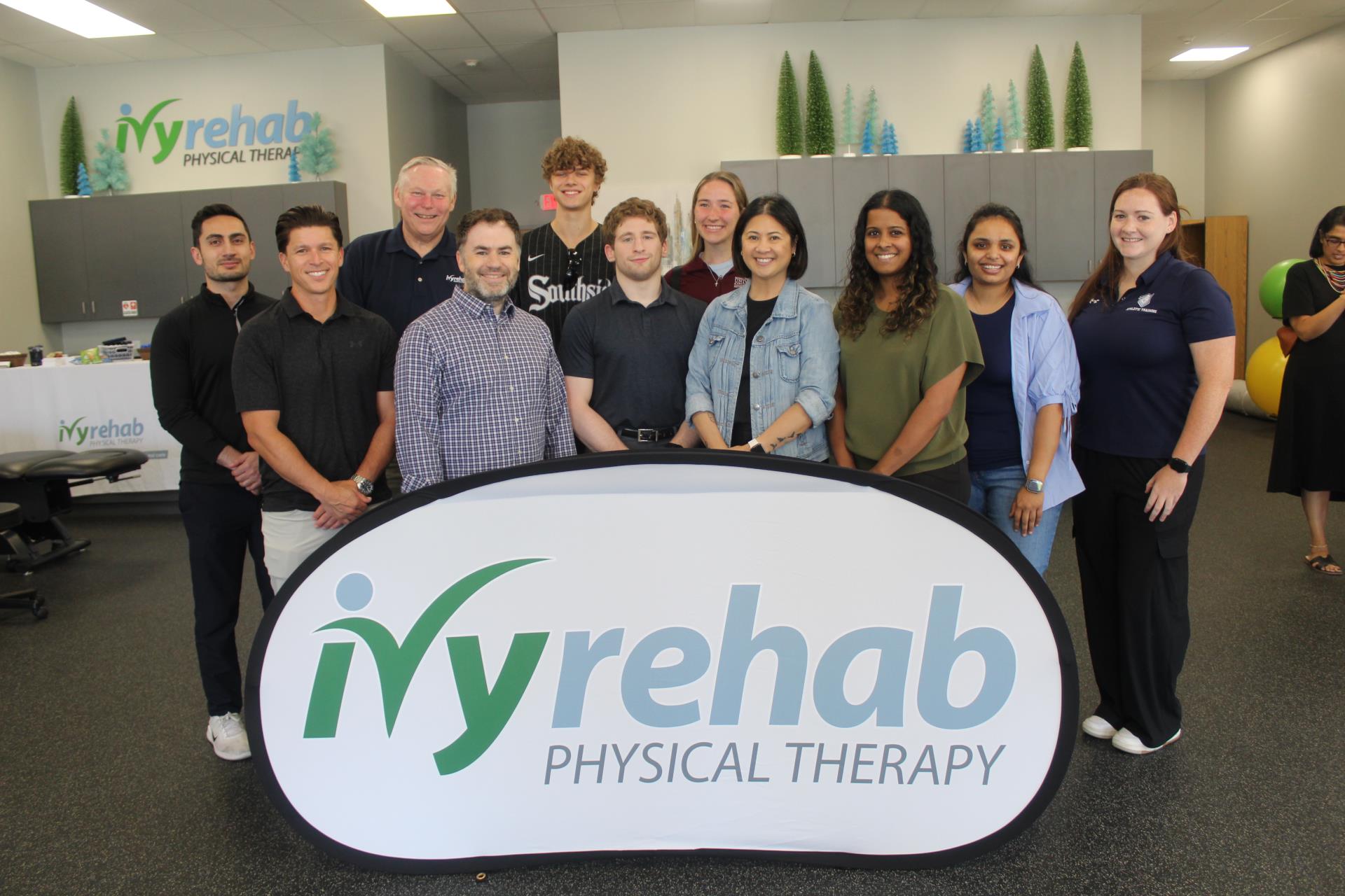 Group of employees lined up smiling at camera. In front is a sign that says ivy rehab physical therapy.