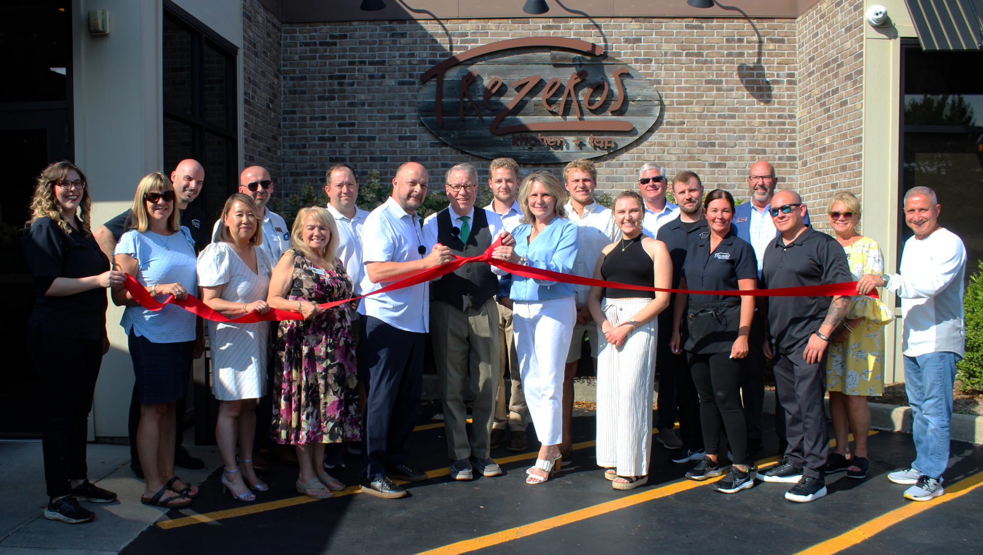 Group of employees, friends, and family lined up with Mayor in front of Trezeros. They are holding a red ribbon tied in the middle.