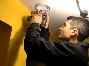 Person with an electric drill installing a smoke alarm on a ceiling.